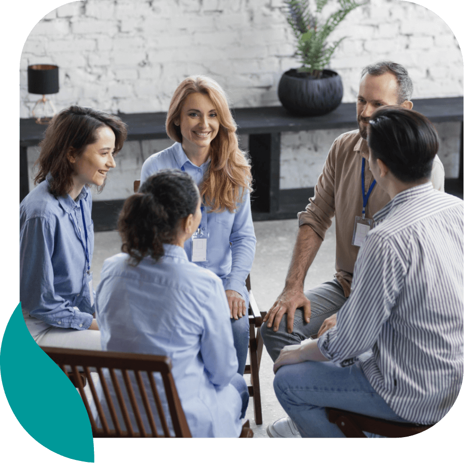 A diverse group of five adults sit in a circle, smiling and talking, in a bright room with a brick wall and plant, suggesting an alcohol rehab in Philadelphia PA or support group focused on healing together.