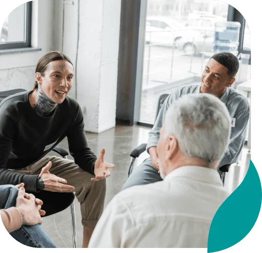 Three people sit in a circle having a lively discussion in a bright, modern room with large windows, sharing their experiences as part of addiction treatment in Philadelphia PA. One person speaks animatedly while the others listen and engage.