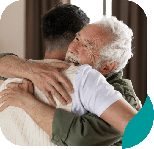 An elderly man with white hair smiles warmly while hugging a younger man indoors. Both appear happy and content, embracing each other closely in a heartfelt moment, perhaps celebrating progress after addiction treatment in Philadelphia PA.