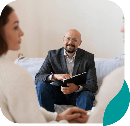 A smiling man with glasses and a notebook sits on a couch, appearing to counsel two people—reflecting the supportive environment offered for depression treatment in Philadelphia PA.
