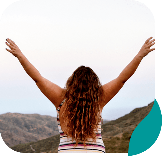 A person with long, wavy hair stands outdoors facing away, arms raised in a V shape, overlooking hills under a pale sky. The person wears a striped sleeveless top.