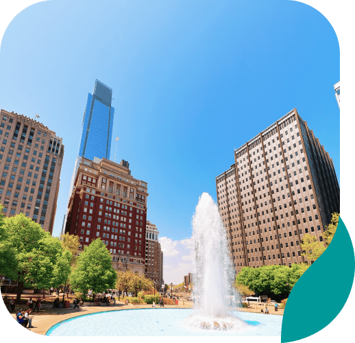A cityscape view featuring a fountain in the foreground, surrounded by green trees and tall buildings under a clear blue sky.