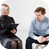 A woman with glasses holds a clipboard and talks to a seated man who looks pensive and has his hands clasped, suggesting a counseling or therapy session in a bright, minimalist room.