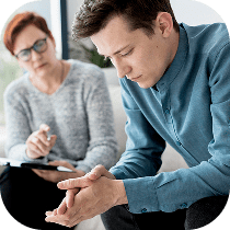 A young man sits with his head down and hands clasped, looking thoughtful or troubled, while a woman with glasses takes notes and listens attentively in a counseling or therapy session.