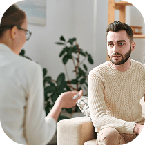 A man in a cream sweater sits on a couch, attentively listening to a woman with glasses who appears to be counseling him in a comfortable, well-lit room with plants in the background.