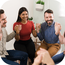 Four people sit in a circle holding hands and smiling, appearing to participate in a supportive or group therapy session in a bright, welcoming room with plants on the wall.