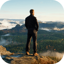 A person stands on a rocky cliff edge, looking out over a vast forested landscape with mountains and mist in the distance under a clear sky.