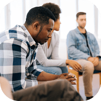A man in a plaid shirt sits pensively in a group therapy session, with two other people seated nearby, all appearing engaged in a serious conversation.