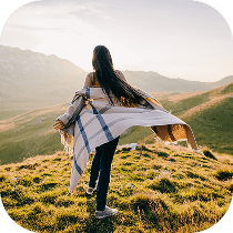 A person with long hair stands on a grassy hilltop, facing away and holding a blanket that flows in the wind, with mountains and a soft sunset in the background.