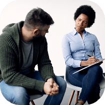 A woman with a notepad listens intently to a man who appears to be speaking to her; both are seated and facing each other in a counseling or therapy setting.