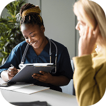 A healthcare professional in navy scrubs and a stethoscope smiles while writing on a clipboard, sitting across from a person in a yellow top who is touching their face.