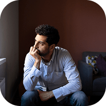 A man with dark hair and a beard sits indoors, resting his chin on his hand and gazing thoughtfully out of a window, with a couch and cushions in the background.