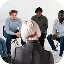 A group of people sits in a circle, attentively listening to a person with short blonde hair who faces them, suggesting a therapy or support group meeting in a bright, minimalist room.