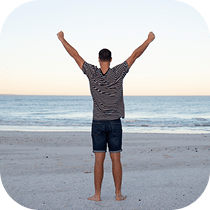 A person stands barefoot on a sandy beach facing the ocean, arms raised in celebration or triumph, wearing a striped shirt and shorts under a clear sky.