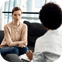 A woman in a light-colored sweater sits on a couch facing a person holding a clipboard, suggesting a counseling or therapy session in a modern, bright office setting.