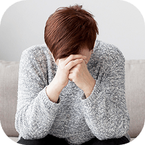 A person with brown hair wearing a gray sweater sits on a couch, leaning forward with their head down and hands clasped over their face, appearing upset or stressed.