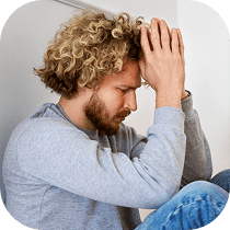 A man with curly blond hair and a beard sits with his knees up, leaning his elbows on them and pressing his hands together against his forehead, appearing stressed or deep in thought.