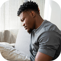 A man sits on a couch indoors, leaning forward with a pained expression, resting his hand on his stomach as if experiencing discomfort.