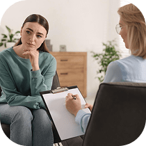 A woman sits with her hand on her chin, looking thoughtful, while another person holding a clipboard appears to be taking notes during a counseling or therapy session in a cozy, well-lit room.