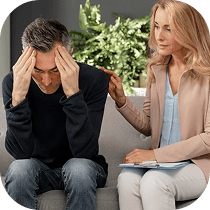A woman sits beside a distressed man on a couch, gently touching his shoulder to comfort him. The man has his elbows on his knees and holds his head in his hands. A plant sits in the background.