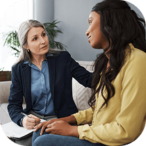 A therapist with gray hair and a blue blazer sits beside a young woman in a yellow shirt, listening attentively and offering support in a cozy, well-lit room.