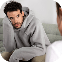 A man wearing a gray hoodie sits on a couch, leaning his head on his hand and looking thoughtfully at another person who is partially visible in the foreground.