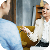A professional woman in a light blazer talks to another person, holding a pen and gesturing with her hand, during what appears to be a counseling or consultation session.