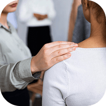 A person gently places a comforting hand on another person's shoulder, offering support. Blurred figures stand in the background, suggesting a group setting.