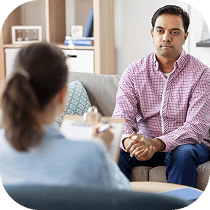A man sits on a couch with his hands clasped, facing a woman who appears to be a therapist taking notes during a counseling session in a comfortable office setting.