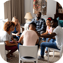 A diverse group of six adults sit in a circle of chairs in a cozy living room, engaging in conversation and offering support to each other.