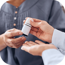 Close-up of two people’s hands as one person dispenses medication or pills from a small white bottle into the other person’s open palm. Both are wearing long-sleeved shirts.
