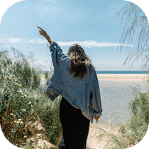 A woman with long hair, wearing a denim jacket and black dress, walks toward a sandy beach with water and blue sky, her arm raised as she points ahead; greenery surrounds her path.