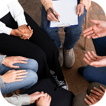 A group of people sitting in a circle, some holding hands and others gesturing, with one person holding a clipboard and pen, suggesting a group discussion or therapy session.