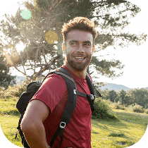 A young man with a beard, wearing a red shirt and a backpack, smiles while standing outdoors in a grassy area with trees and sunlight in the background.