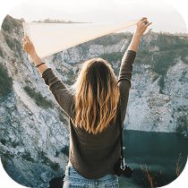 A person with long, blonde hair stands outdoors facing a rocky cliff, holding a white cloth above their head. They wear a brown sweater and jeans, with a camera hanging at their side.