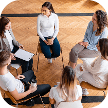 Five people sit in a circle on chairs, engaged in conversation or discussion, in a bright room with wooden flooring. One person is holding a clipboard, another has a coffee cup.
