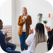 A woman stands and smiles while a group of people sitting in a semi-circle clap for her in a bright, modern room with plants and shelves.