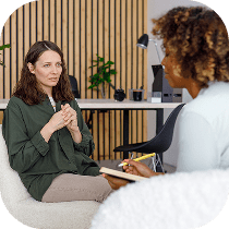 A woman with brown hair sits on a chair, speaking with another woman who is writing in a notebook during a therapy or counseling session in a modern office.