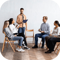 A group of five people sit in a circle on wooden chairs having a discussion, with one man standing and holding a clipboard, while others listen and take notes.