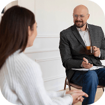 A man sitting on a chair holding a cup and clipboard, smiling and talking with a woman who is facing him. They appear to be in a casual, indoor setting, possibly having a conversation or counseling session.