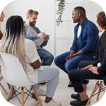 Four people sit in a circle, engaged in conversation. One person is holding papers and appears to be leading the discussion. The setting is a bright indoor space with white chairs and a plant in the background.