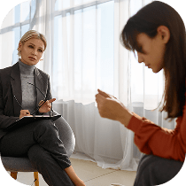 A therapist sits with a notepad, attentively listening to a woman who appears distressed during a counseling session in a bright, softly lit room.
