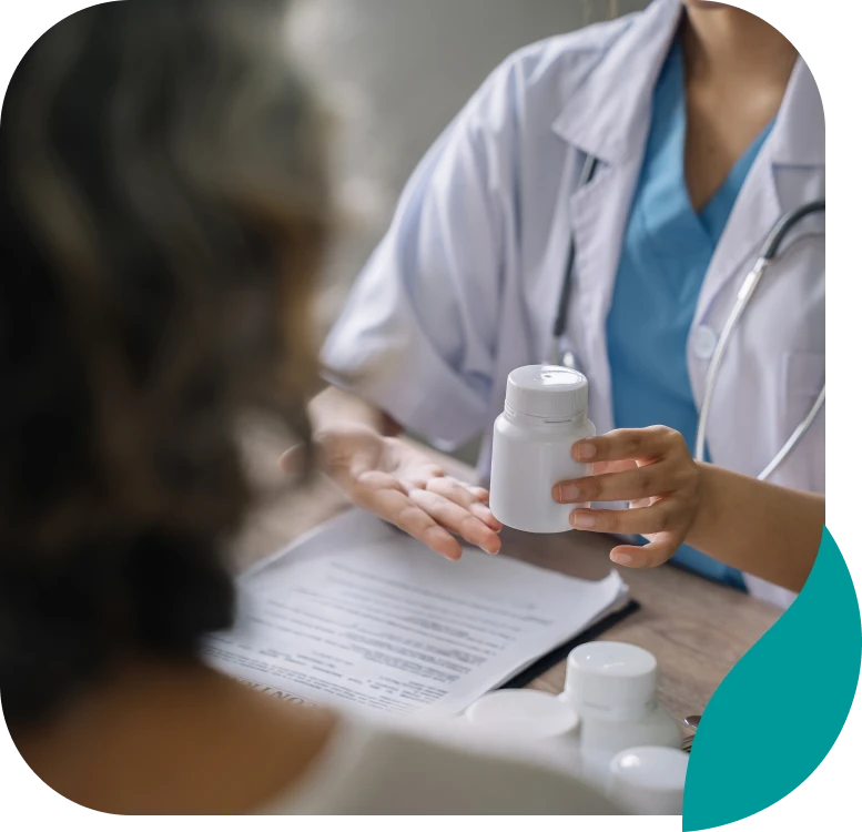 A doctor in a white coat and blue scrubs sits at a desk, holding a white medicine bottle while talking to a patient. Medical documents and other bottles are on the table.