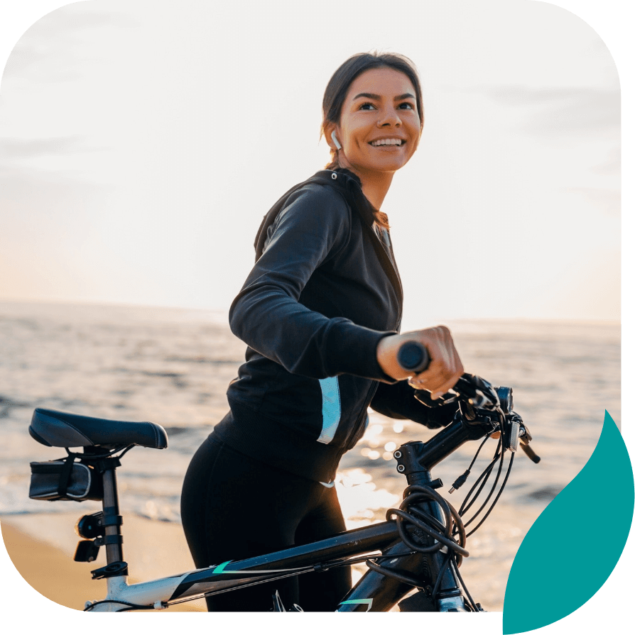 A smiling woman in athletic wear stands beside her bicycle on a sandy beach at sunset, looking into the distance. The ocean and sky create a peaceful background.