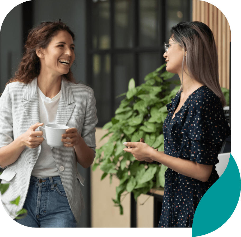 Two women standing and smiling while having a conversation; one is holding a white mug, and there are green plants in the background.