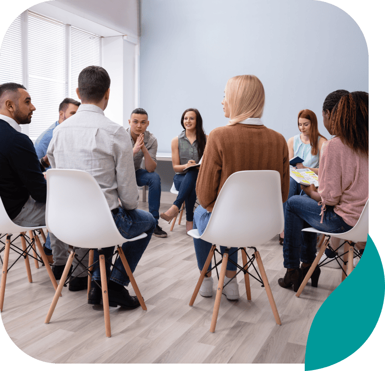 A diverse group of people sits in a circle on white chairs in a bright room, engaging in a group discussion or support meeting.