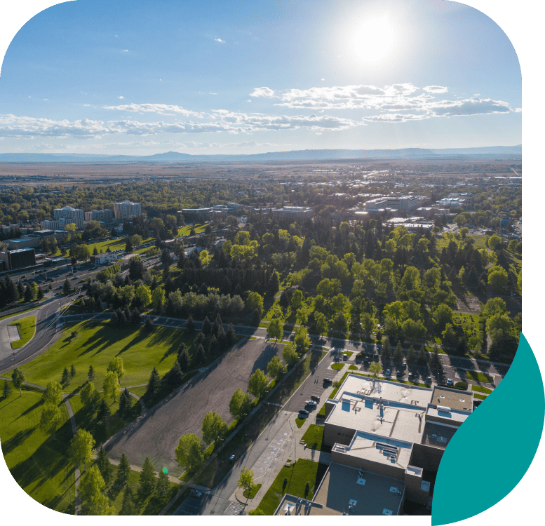 Aerial view of a sunlit cityscape with many green trees, open grassy areas, and buildings. The sky is mostly clear with some clouds, and the sun is shining brightly.