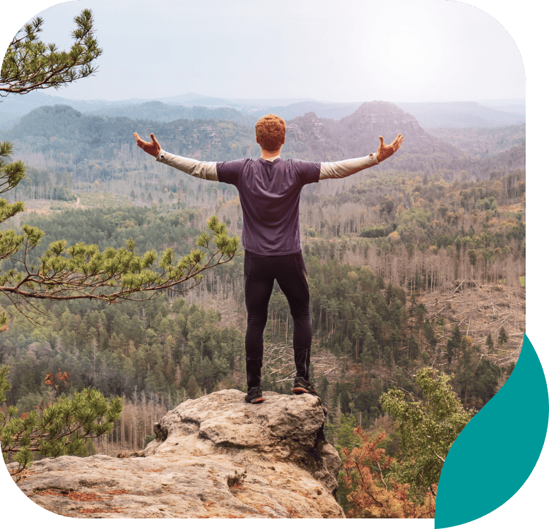 A person with outstretched arms stands on a rocky cliff, overlooking a vast forested valley and distant mountains under a hazy sky.